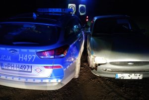 Zdjęcie zrobione w porze wieczorowo-nocnej, widnieje na nim policyjny radiowóz oznakowany sfotografowany z tyłu. Równolegle do radiowozu stoi uszkodzony szary samochód.