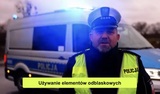 Zdjęcie ze spotu załączonego do artykułu. Zdjęcie przedstawia policjanta ruchu drogowego na tle radiowozu policyjnego. W dolnej części żółty pasek z napisem Używanie elementów odblaskowych.