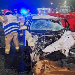 na zdjęciu widnieje biały samochód z rozbitym przodem, wokół pojazdy uprzywilejowane z lampami błyskowymi oraz straż pożarna