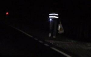Na zdjęciu znajduje się sylwetka pieszego poruszającego się droga w całkowitych ciemnościach. Pieszy ma na sobie kamizelkę, na której znajdują się umieszczone poziomo odblaskowe pasy. Kamizelka jest widoczna w światłach samochodu.