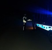 Zdjęcie przedstawia policjanta, który stoi obok radiowozu w ciemnosinych i trzyma elementy odblaskowe demonstrując jak odbijają światło