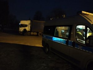 zdjęcie zrobione zostało w porze wieczorowo-nocnej. Na ciemnym parkingu stoi policyjny radiowóz a obok kontrolowana ciężarówka w jasnym kolorze.