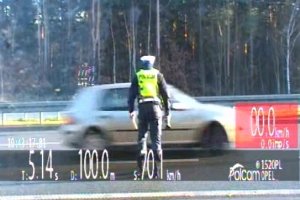Na zdjęciu widać umundurowanego policjanta w kamizelce odblaskowej, który stoi tuz przy drodze i patrzy na samochód w kolorze szarym jadący z dużą prędkością.