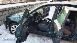 Zdjęcie przedstawia samochód osobowy w kolorze zielonym, który przodem uderzył w niski murek. Drzwi do samochodu są otwarte i nie ma w nim kierowcy ani pasażera. Zdjęcie zrobione zostało w dzień, na drodze leży śnieg.