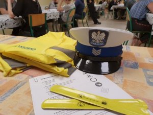 na rysunku widać policyjna czapkę, która leży na stole w pomieszczeniu a obok pookładane są elementy odblaskowe, opaski w kolorze żółtym i kamizelki