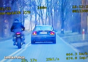 Zdjęcie jest &quot;stop klatką&quot; z policyjnego wideorejestratora. Jest ono w tonacji niebieskiej. Widać na nim samochód osobowi i motocyklistę jadących w tym samym kierunku na dwóch sąsiadujących ze sobą pasach ruchu. Zrobione zostało na drodze położonej wśród drzew.
