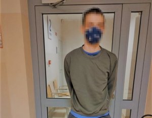 Na zdjęciu widać zatrzymanego mężczyznę, znajdującego się w budynku policji, ubranego w bluzę w kolorze oliwkowym, z maseczka na twarzy.