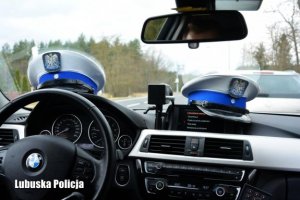 Zdjęcie przedstawia zbliżenie wnętrza radiowozu marki BMW, widać tzw. kokpit czyli liczniki samochodu, kierownice, a także położone we wnętrzu czapki policjantów ruchu drogowego.