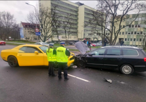 Na zdjęciu widać dwa samochody, jeden w kolorze żółtym, drugi czarnym, które zderzyły się czołowo, obok samochodów swoi dwója umundurowanych policjantów w żółtych kamizelkach z napisem Policja, którzy dokonują oględzin kolizji.
