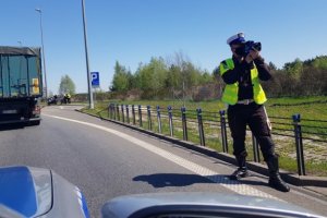 Policjant umundurowany stoi na poboczu drogi i używa wideorejestratora do mierzenia prędkości pojazdów, trzyma go na wysokości oczu