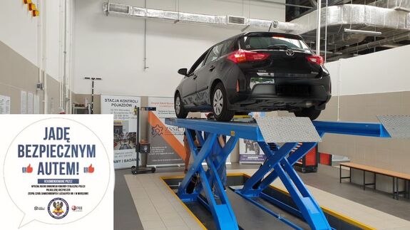 Samochód osobowy, koloru czarnego, jest podniesiony na podnośniku warsztatowym. Z lewej strony znajduje się logo akcji Jadę bezpiecznym autem.