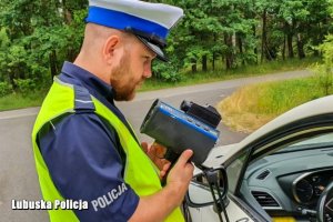 Na pierwszym planie stoi policjant w białej czapce i kamizelce odblaskowej, w ręku trzyma urządzenie koloru niebieskiego do pomiaru prędkości pojazdów, w prawym rogu widać fragment pojazdu.
