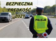 Policjant ruchu drogowego obserwuje ruch drogowy stojąc w pobliżu drogi, na zdjęciu widnieje napis &quot;bezpieczna prędkość 2021&quot;