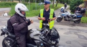 Umundurowany policjant ruchu drogowego legitymuje motocyklistę zatrzymanego do kontroli, motocyklista porusza się czarnym motocyklem i jest w czarnym stroju. W pobliżu stoi policyjny, oznakowany motocykl.