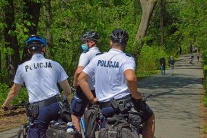 trzech policjantów na rowerach pełnią służbę na drodze , każdy jest w białej podkoszulce z napisem Policja