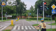 Miasteczko ruchu drogowego z wydzielonymi ulicami, znakami drogowymi i przejściami dla pieszych, na kto rym widać dziewczynkę w kasku rowerowym przygotowującą się do jazdy.