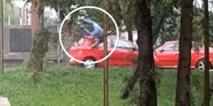 Jadący samochód w kolorze czerwonym, na masce kierujący hulajnogą. Zdjęcie jest stop-klatka z filmu zamieszczonego poniżej.