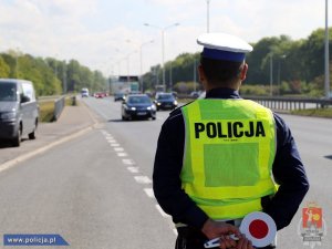policjant ruchu drogowego stoi przy ciągu komunikacyjnym ma na sobie żółtą kamizelkę z napisem POLICJA, w ręku trzyma tarczę do zatrzymywania pojazdów.