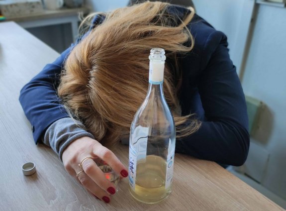 Zdjęcie przedstawia kobietę pochyloną nad stolikiem, na którym stoi butelka z alkoholem