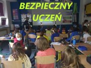 Grupa dzieci w klasie, podczas spotkania z policjantem, na zdjęciu napis w jaskrawożółtym kolorze: Bezpieczny Uczeń