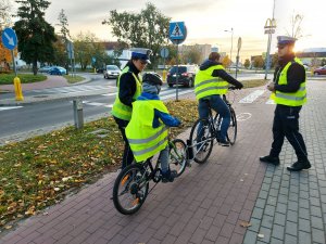 Na ścieżce rowerowej widać dwoje policjantów i dwóch młodych rowerzystów. Wszyscy noszą na sobie kamizelki odblaskowe.