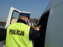 Umundurowany policjant ruchu drogowego w odblaskowej kamizelce z napisem POLICJA kontroluje kierowce białego busa.