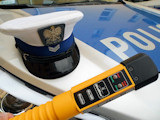 Zaimprowizowane zdjęcie, na którym na masce policyjnego radiowozu lezy czapka policjanta ruchu drogowego i urządzenie do wykrywania alkoholu w wdychanym powietrzu w kolorze żółtym.