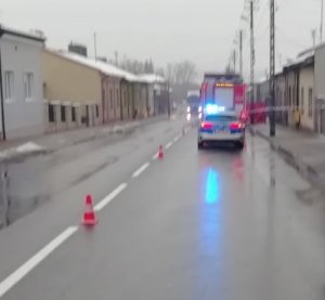 Wąska ulica zabudowana, na której z oddali widać stojący radiowóz policyjny i samochód straży pożarnej. jezdnia jest mokra.
