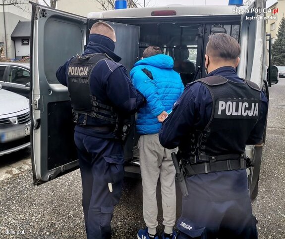 Na zdjęciu widać dwóch umundurowanych policjantów, obróconych plecami, przed nimi stoi mężczyzna w niebieskiej kurtce i szarych spodniach. Mężczyzna ma ręce z tyłu pleców. Przed nimi stoi radiowóz policyjny koloru szarego.