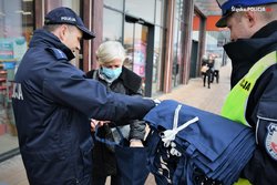 dwóch policjantów na ulicy miasta wręcza starszej kobiecie granatowa torbę z elementem odblaskowym