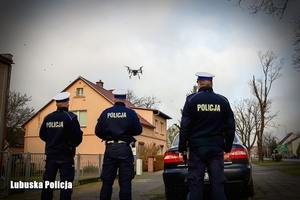 trzej policjanci widoczni od tyłu w kurtkach z napisem POLICJA stoją przy drodze, na niebie widać latający dron