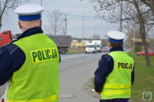 dwóch policjantów w kamizelkach odblaskowych z napisem POLICJA stoi przy ruchliwej drodze
