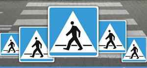 Z tyłu zdjęcia znajduje się przejście dla pieszych, na pierwszym planie pięć znaków drogowych - każdy z niebieskim tłem, białym trójkątem na środku, i grafiką przedstawiającą idącego człowieka.