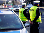 dwóch policjantów ruchu drogowego w kamizelkach odblaskowych z napisem Policja stoi obok radiowozu przy ruchliwej drodze