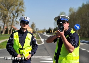 Na zdjęciu widać dwóch policjantów ruchu drogowego, którzy stoją na drodze i kontrolują prędkość pojazdów.