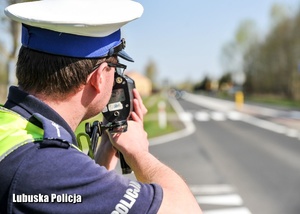 Na zdjęciu widać policjanta w białej czapce oznaczającej policjanta ruchu drogowego, który trzyma urządzenie do pomiaru prędkości.