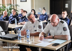 Na pierwszym planie widać policjantów niemieckich w białych koszulach siedzących przy biurku, za nimi siedzą policjanci ubrani w granatowe mundury.