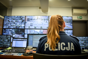 Na pierwszym planie widać policjantkę ubraną w niebieski mundur z napisem POLICJA. przed nią znajduje ściana pokryta monitorami, telewizorami.