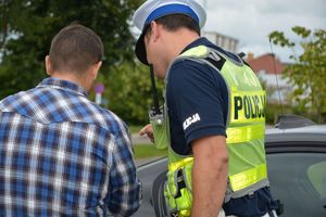 Po lewej stronie widać osobę w  koszuli w kratkę stojącą obok policjanta ubranego w kamizelkę odblaskową z napisem policja