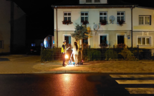 zdjęcie w scenerii wieczorowo-nocnej na oświetlonej ulicy. Policjant rozmawia z kobietą, która stoi przy hulajnodze elektrycznej.