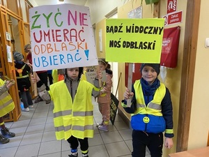 dzieci w kamizelkach odblaskowych i transparentach z napisami zachęcającymi do noszenia elementów odblaskowych