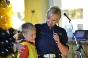 policjantka z mikrofonem stoi obok chłopca, który ma na sobie odblaskowa kamizelkę
