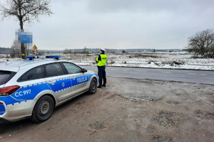 Radiowóz policyjny, przed nim stoi policjant ruchu drogowego, w oddali widać pole przykryte śniegiem.