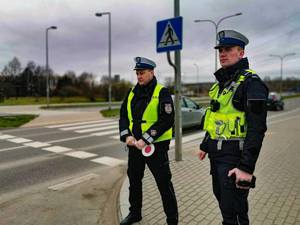 Na pierwszym planie stoją policjanci ruchu drogowego, za nimi widać przejście dla pieszych oraz jadące pojazdy.