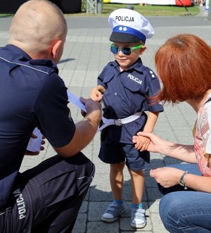 Policjant wręczający kartkę małemu chłopcu, który jest ubrany w strój policjanta. Obok chłopca kuca kobieta i trzyma go za rękę